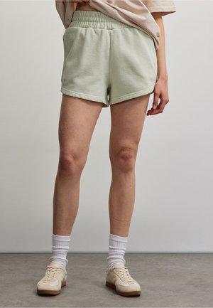 Person, die hellgrüne Shorts mit elastischem Bund, weiße Sportsocken und beigen Sneaker trägt, steht vor einem einfarbigen hellen Hintergrund.