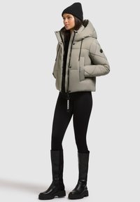 Kvinde iført en lys grå pufferjakke, sort strikket beanie, sorte leggings og sorte knæhøje støvler, stående med hænderne i lommerne.