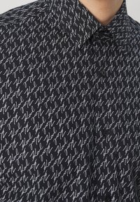 Camicia nera con bottoni, caratterizzata da testo bianco in corsivo su un motivo ripetuto. Il tessuto ha una texture liscia con un design classico del colletto.