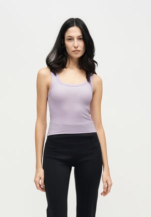 Une femme aux longs cheveux foncés porte un haut sans manches lilas et un pantalon noir sur un fond clair uni.