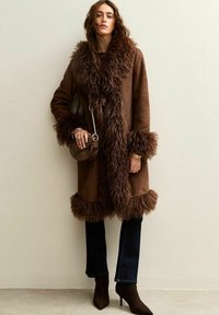 Bruine suède jas met een fluffy bontafwerking bij de kraag en manchetten, gecombineerd met donkere jeans en bruin hoge hakken, terwijl ze een kleine handtas vasthoudt.