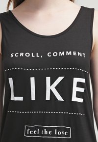 Svart ärmlös bomullstopp med vit tryckt text som säger "SCROLLA, KOMMENTERA GILLA" och "känn kärleken" med streckade linjer.