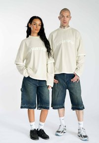Helle beige übergroße T-Shirts mit dem Text "MULTIPLY", kombiniert mit dunklen Denim-Shorts, weißen Socken und Sneakers. Lässiger, entspannter Schnitt.
