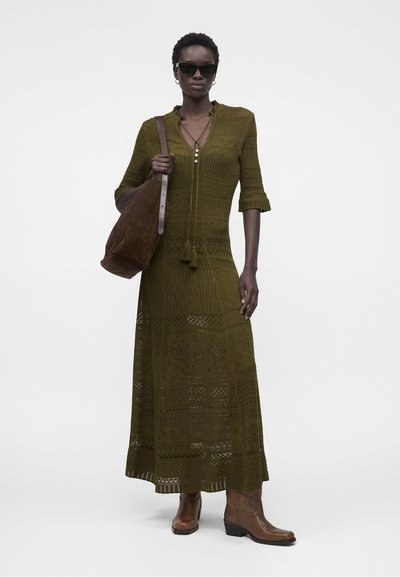 Γυναίκα που φοράει μακρύ φόρεμα με μοτίβο σε ελαιοπράσινο χρώμα, καφέ μπότες, γυαλιά ηλίου και μία μεγάλη καφέ τσάντα ώμου σε απλό φόντο.