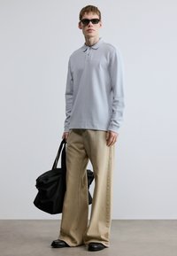 Ανοιχτό γκρι μακρυμάνικο πόλο με απαλή υφή, σε συνδυασμό με φαρδιά, μπεζ παντελόνια και μαύρη τσάντα. Το μοντέλο φοράει σκούρα γυαλιά ηλίου.