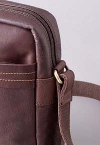 Brown leather bag featuring a zip closure, prominent stitching, a side pocket, and a fabric strap with metal hardware accents.