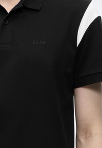Polo noir à manches courtes avec une bande blanche sur l'épaule et « BOSS » brodé sur la poitrine.
