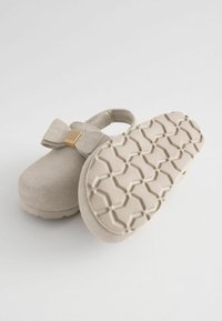 Miękkie beżowe klapki dla malucha z kokardą i złotym detalem, z jednym pokazującym chropowatą, wzorzystą podeszwę dla lepszej przyczepności.