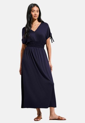 Vrouw met lang zwart haar, gekleed in een marineblauwe midi-jurk met V-hals, korte mouwen en een geplooide taille, staand op blote voeten tegen een witte achtergrond.