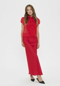 Czerwona bluzka z falbanami w warstwy zestawiona z pasującą długą spódnicą. Gładka tekstura materiału, dopasowany krój, krótkie bufiaste rękawy i czarne szpilki.