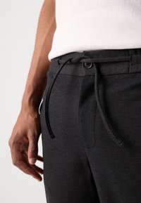 Pantalons de jogging gris foncé avec une taille à cordon de serrage, présentant une fermeture à bouton et une texture lisse, exhibés aux côtés d'une main.