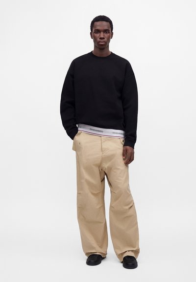 Homme debout portant un sweat-shirt noir, un pantalon beige à jambes larges, des chaussures noires et une ceinture blanche de marque visible au-dessus du pantalon.