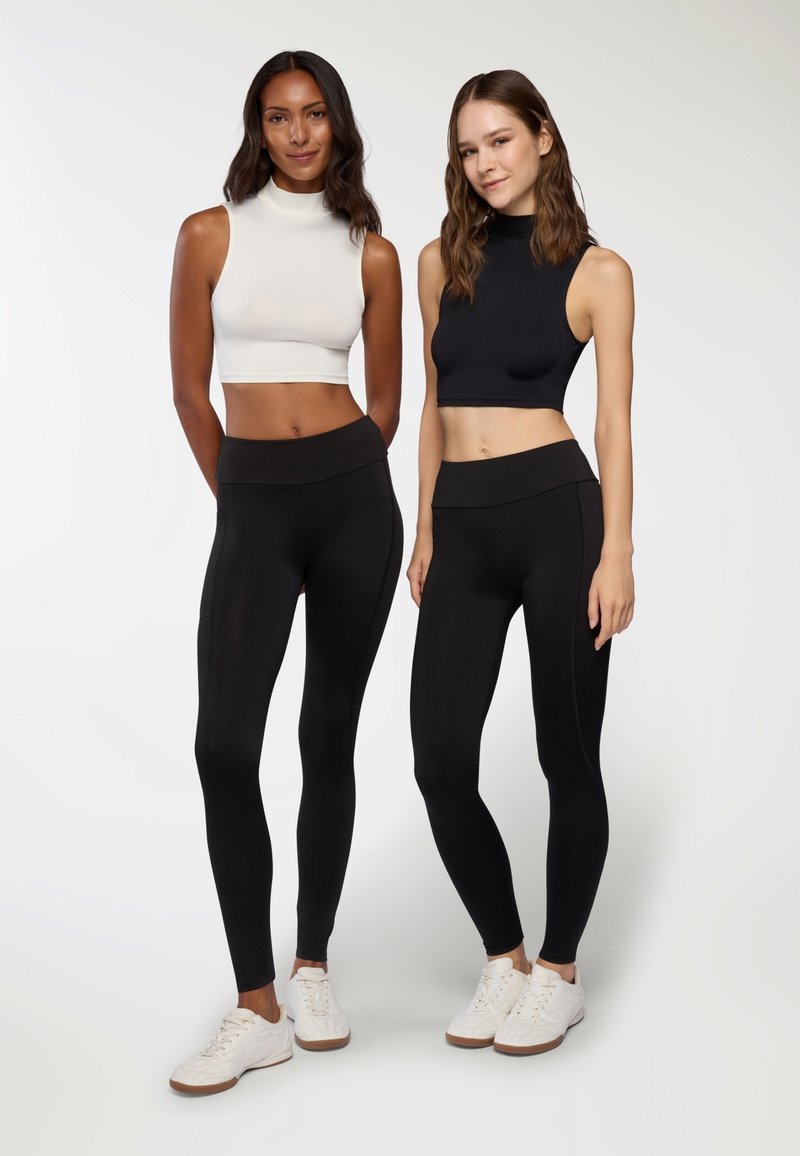Dwie modelki w dopasowanych czarnych legginsach i bezrękawnikach; jeden top jest biały, a drugi czarny. Obie noszą białe sneakersy.
