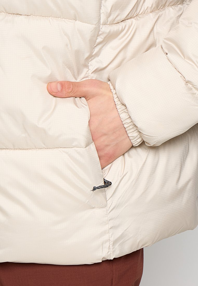 Chaqueta acolchada beige claro con puños acanalados, costuras visibles y un bolsillo lateral con cremallera, que muestra una textura suave y brillante.
