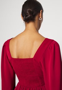 Rotes Kleid mit quadratischem Rückenteil, gerafftem Oberteil und Puffärmeln. Glatte Satintextur mit tailliertem Schnitt und ausgestellt Rock.