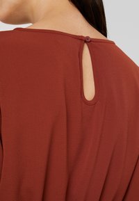 Gros plan sur le dos d'un haut couleur rouille avec un tissu texturé, une ouverture en forme de trou de serrure et une seule fermeture à bouton près du col.