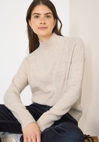 Beige neule, jossa on korkea kaulus ja teksturoitu kuvio. Tiukkataskuiset hihat ja rento malli. Puettuna tummien farkkujen kanssa, istuu pinnalla.