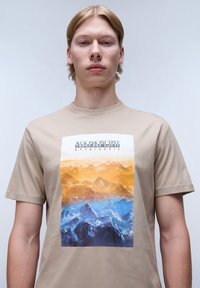 Beige bavlněné tričko s grafikou hory v oranžových a modrých odstínech, s textem "NAPAPIJRI" nahoře a kulatým výstřihem.