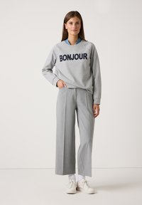 Frau steht in grauem "BONJOUR" Sweatshirt, hellgrauen weit geschnittenen Hosen, weißen Socken und weißen Sneakers vor einem einfarbigen Hintergrund.