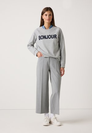 Frau steht in grauem "BONJOUR" Sweatshirt, hellgrauen weit geschnittenen Hosen, weißen Socken und weißen Sneakers vor einem einfarbigen Hintergrund.