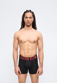 Άνδρας χωρίς πουκάμισο με μακριά dreadlocks, φορώντας μαύρα εσώρουχα Calvin Klein boxer briefs, στέκεται μπροστά σε ανοιχτό γκρι φόντο.