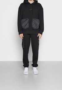 Person som bär en svart huvtröja med stora framfickor, matchande svarta byxor med sidofickor, och vita sneakers, stående mot en enfärgad bakgrund.