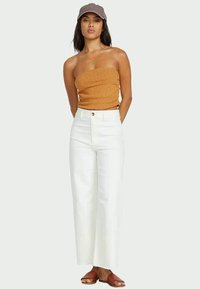 Sans bretelles, haut strié en moutarde, pantalon large taille haute blanc avec bouton sur le devant, et sandales plates marron. Style décontracté et ajustement relax.