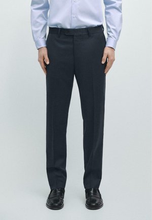 Ein Mann trägt schwarze Anzughose, schwarze Leder-Loafer und ein hellblaues langärmliges Hemd und steht mit den Händen an den Seiten.