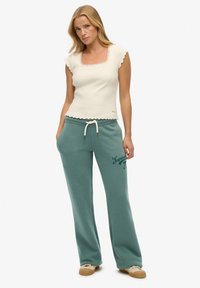 Crème ribbeltop met geschulpte randen, gecombineerd met teal trainingsbroek met een trekkoord en "Superdry 73" in het groen gedrukt.