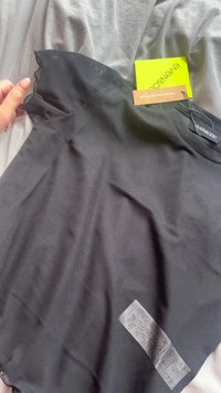 Černé tričko s krátkým rukávem a zaobleným výstřihem, vyrobené z měkkého materiálu, s etiketou pro recyklovaný polyester. Jsou připojené štítky.
