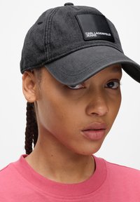Černá denimová baseballová čepice s plochým kšiltem. Obsahuje obdélníkovou černou koženou nášivku s nápisem "KARL LAGERFELD JEANS." Texturovaná tkanina.