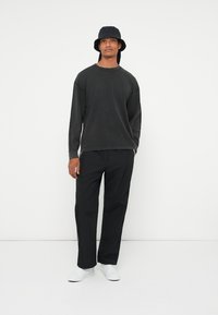 Crna majica dugih rukava s okruglim izrezom, uparena s crnim širokim hlačama i bijelim tenisicama. Crni šešir u obliku kante upotpunjuje odjevnu kombinaciju.