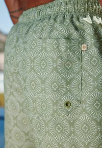 Zöld mintás fürdőnadrág fehér geometriai mintákkal, rugalmas derékpánttal, oldalsó zsebbel és egy kis napocska logó részlettel.