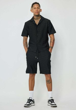 Chemise noire à manches courtes avec un motif en diamant texturé, associée à un short noir assorti. Elle est accompagnée de baskets et de chaussettes blanches mi-mollet.