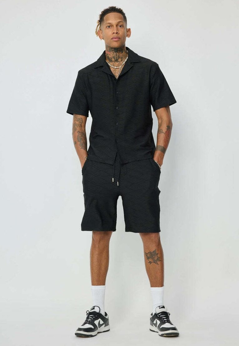 Chemise noire à manches courtes avec un motif en diamant texturé, associée à un short noir assorti. Elle est accompagnée de baskets et de chaussettes blanches mi-mollet.