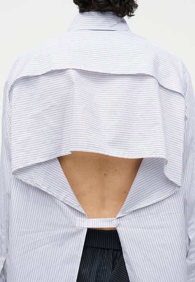 Person mit kurzen dunklen Haaren, der ein helles, hinten offenes Hemd mit Nadelstreifen trägt, das die obere Rückenhaut freilegt, kombiniert mit einer dunklen Nadelstreifenhose.