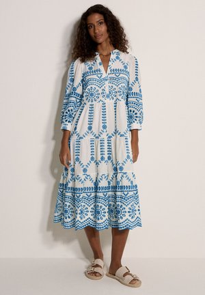 Donna con un vestito midi bianco con ricamo floreale blu e maniche a sbuffo, abbinato a sandali beige, in piedi contro un muro semplice.