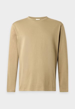 Junger Mann trägt beige Langarmshirt, blaue Jeans, schwarz-weiße Sneakers, mit einem karierten Hemd um die Taille gebunden vor einfarbigem Hintergrund.