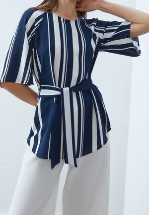 Vrouw draagt een marineblauw-wit verticaal gestreept topje met ellebooglange mouwen en een bijpassende tailleband, gecombineerd met witte broek met wijde pijpen.