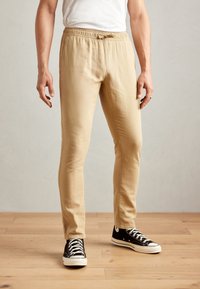 Beige, lightweight, elastic waist pants with a drawstring, featuring a slim fit and paired with black low-top sneakers with white soles.