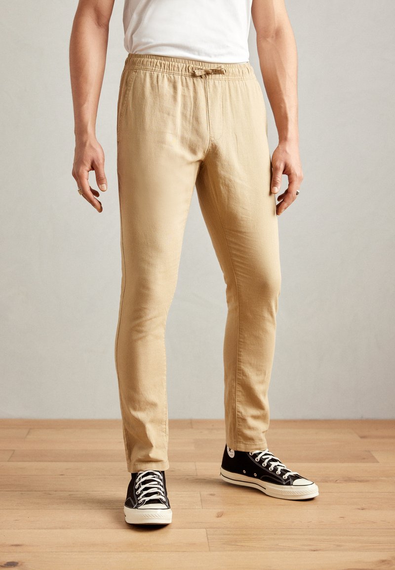 Beige, lightweight, elastic waist pants with a drawstring, featuring a slim fit and paired with black low-top sneakers with white soles.