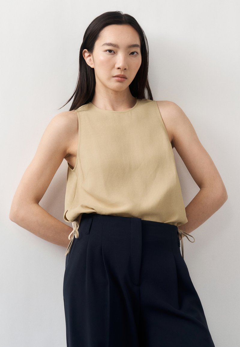 Femme aux cheveux raides et foncés, vêtue d'un haut beige sans manches rentré dans un pantalon noir taille haute, debout devant un fond clair uni.