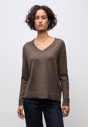 Jersey de color marrón con cuello en V en un tejido suave, corte holgado, con puños y dobladillo acanalados, combinado con unos jeans azul oscuro, presentando un diseño simple y limpio.