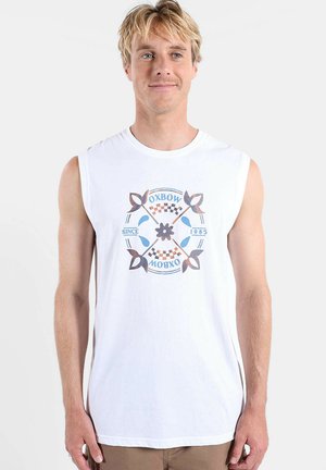 Homme blond portant un débardeur blanc avec le logo Oxbow bleu et marron et un motif floral, se tenant devant un fond blanc uni.