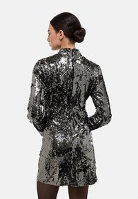 Robe noire et argentée à paillettes avec un col haut, des manches longues et une coupe ajustée, dotée d'une fermeture éclair au dos. Surface texturée.