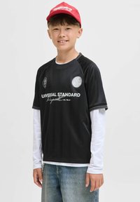 Svart idrottströja med vertikala ränder, broderade logotyper och vit text. Bärs över en vit långärmad tröja. Kombinerad med ljusa jeans.