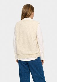 Krämfärgad stickad väst med ribbad nederkant, buren över en vit skjorta, med en avslappnad passform och synlig textur.