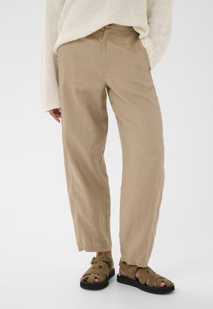 Osoba ubrana w beżowe spodnie o szerokich nogawkach, kremowy sweter z dzianiny oraz brązowe sandały zamszowe z czarną podeszwą na jednolitym białym tle.