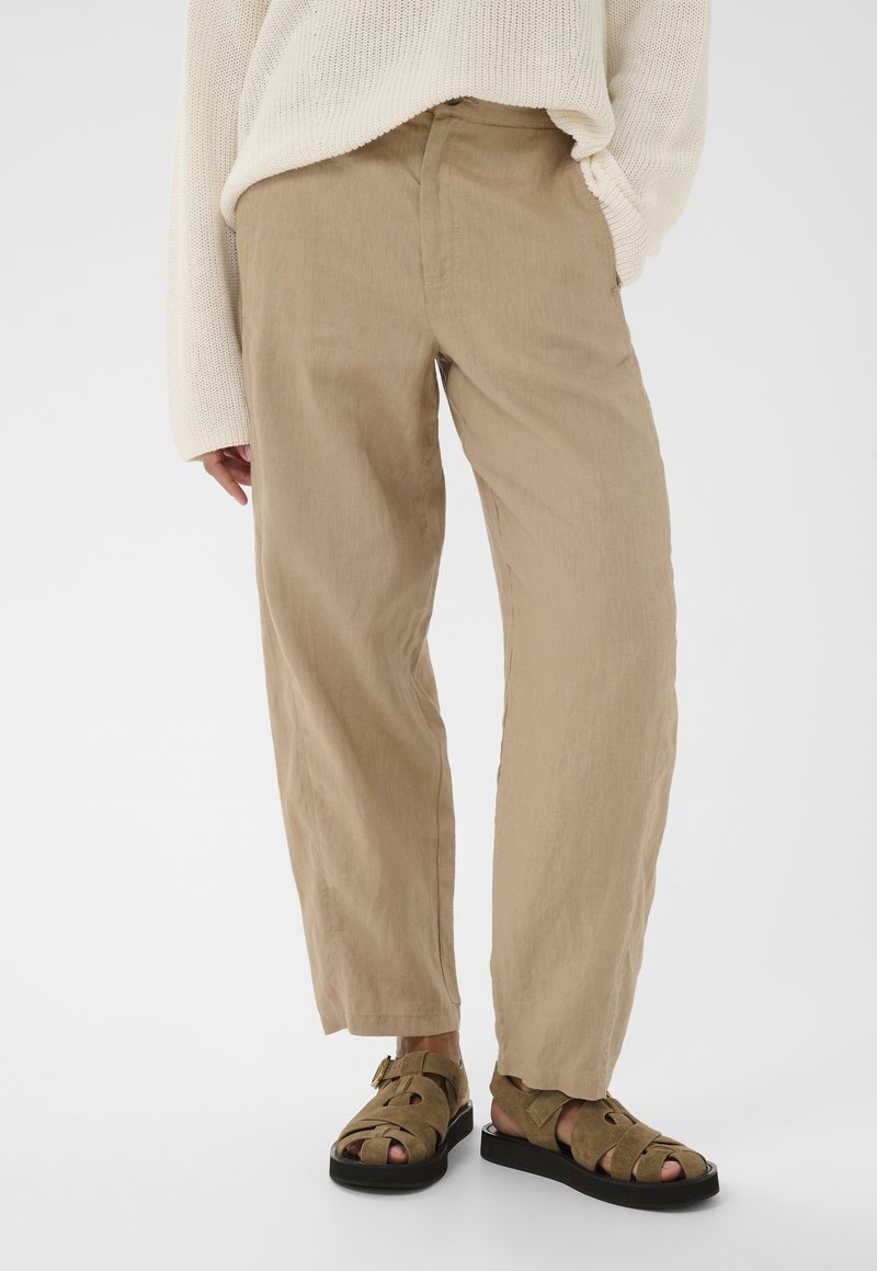 Personne portant un pantalon beige à jambes larges, un pull en maille crème et des sandales en daim marron avec des semelles noires sur un fond blanc uni.