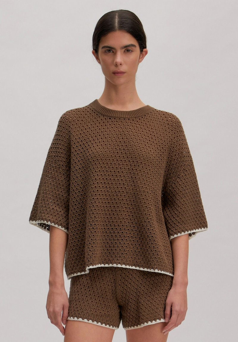 Woman wearing brown crochet knit short-sleeve top and matching shorts with white trim, standing against a plain background.